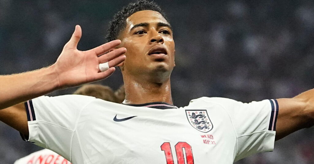 Footballer Jude Bellingham spreading his arms and looking upwards during the World Cup 2026 while wearing the England national team football kit of a white shirt with blue trim and his number in red on the shirt with dark blue shorts and white trim featuring the England badge and his number in white