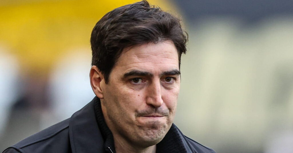 Football manager Andoni Iraola looking to his left while pursing his lips in an outdoor setting. The coach is wearing a dark jacket