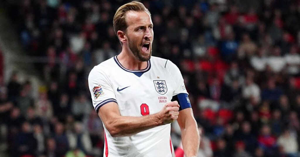 Footballer Harry Kane celebrating a goal by raising his right arm and clenching his fist while roaring and looking to his left while the player for Bundesliga team Bayern Munich Football Club is playing in the England national football team home shirt of white with dark blue details with the Nike logo and Three Lions crest on the chest above his number nine in red with a crowd blurrily visible behind him.