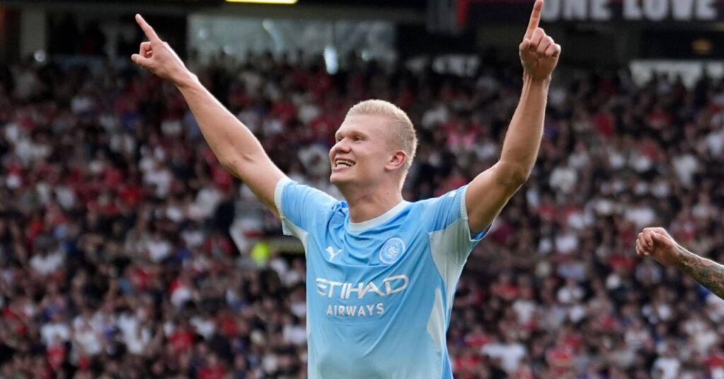 Norway footballer Erling Haaland raising both arms and smiling to celebrate a goal in a stadium while playing for Premier League team Manchester City Football Club in a light blue and white shirt in a stadium
