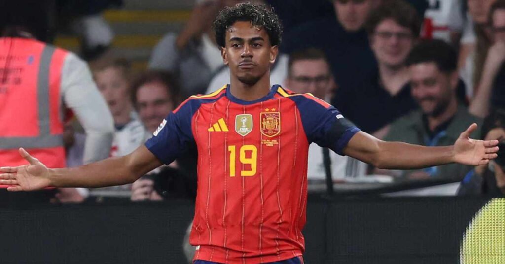 Barcelona footballer Lamine Yamal lifting both arms while on a football pitch wearing the Spain national team football shirt of red, blue and yellow
