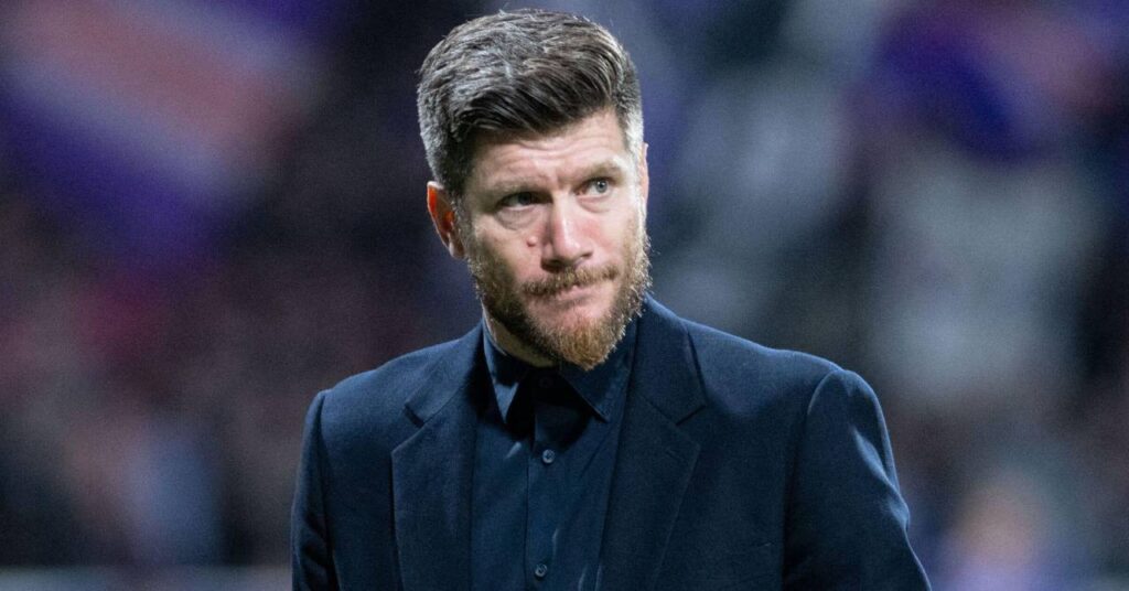 Football manager and Scotland national team head coach Sebastien Pocognoli looking to his left on a pitch in a stadium while wearing a dark suit jacket and shirt