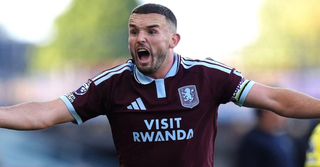 Scotland footballer John McGinn shouting and raising both arms while playing for Premier League team Aston Villa Football Club in a claret home shirt with light blue trim and the Premier League lion logo on the right arm and the words Visit Rwanda and the Adidas and Aston Villa crest on the chest and a captain armband on his left arm.