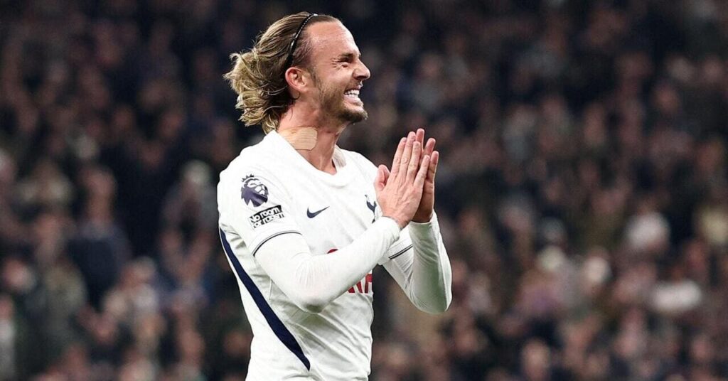 Footballer James Maddison pushes his raised hands together and smiles while looking to his left to celebrate a goal for Premier League team Tottenham Hotspur Football Club in the white Spurs home shirt with the Nike tick logo and club cockerel badge visible on his chest in dark blue.